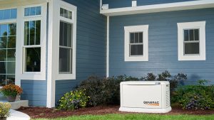 Standby Generac home backup generator installed beside a blue house with white trim, surrounded by landscaped shrubs and flowers.